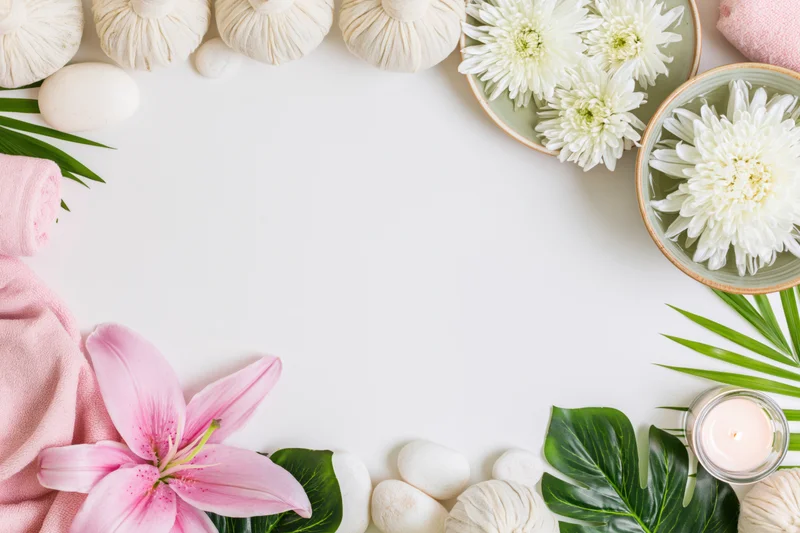 Spa wellness flat lay arranged as a top-and-bottom border with flowers, towels, stones, and leaves on white surface