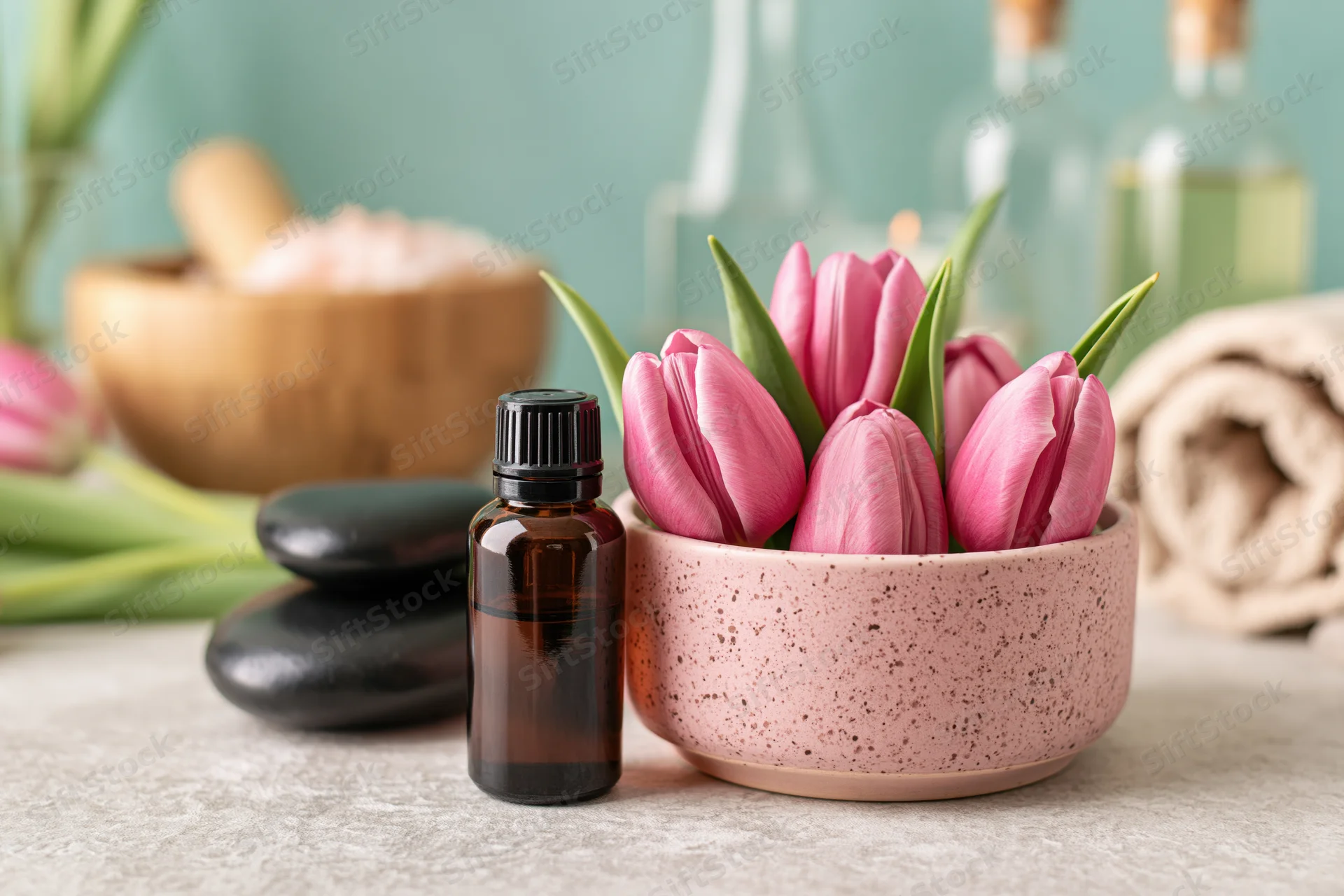 Essential oil bottle and pink tulips in a speckled bowl on a light grey surface with spa stones, towel, mortar and pestle in background