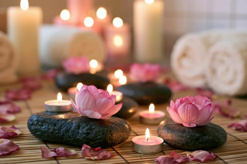 Pink lotus flowers and lit candles arranged on smooth dark stones on a bamboo mat with scattered petals and rolled towels in the background