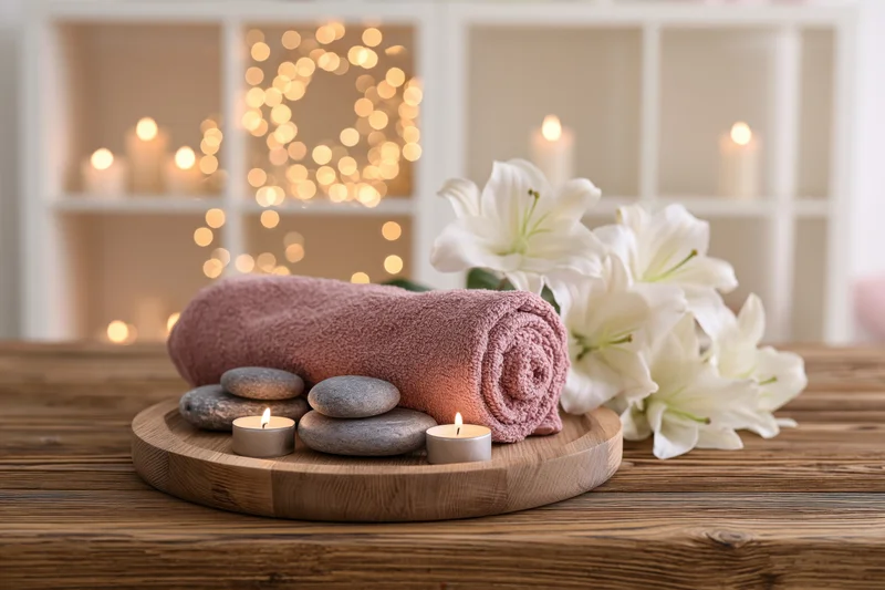 Rolled towel and smooth stones with lit candles and white lilies on wooden tray on table