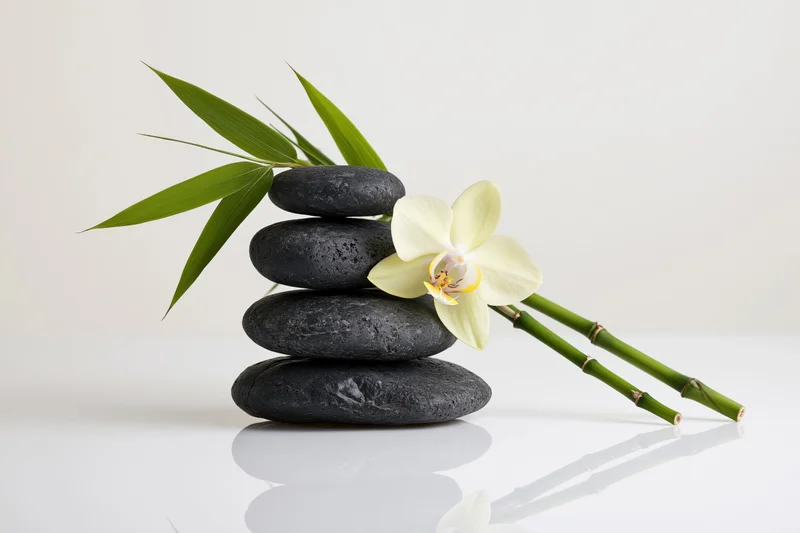 Stack of smooth dark grey basalt stones with bamboo and white orchid on reflective white surface