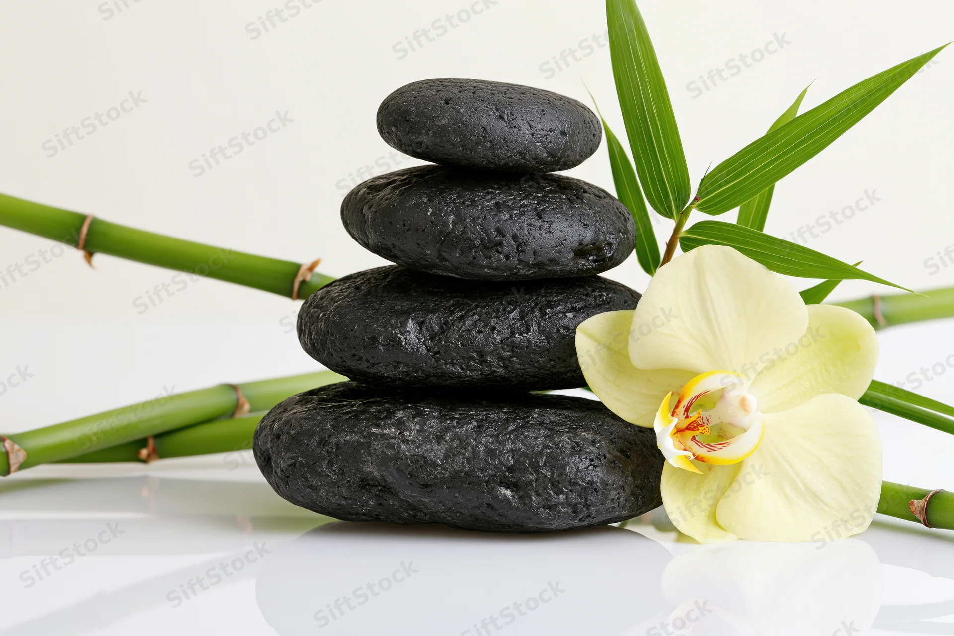 Stack of smooth dark grey basalt stones balanced with green bamboo and a white orchid on a reflective white surface