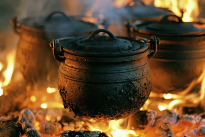 Three blackened cast iron potjie pots with lids cooking over an open fire