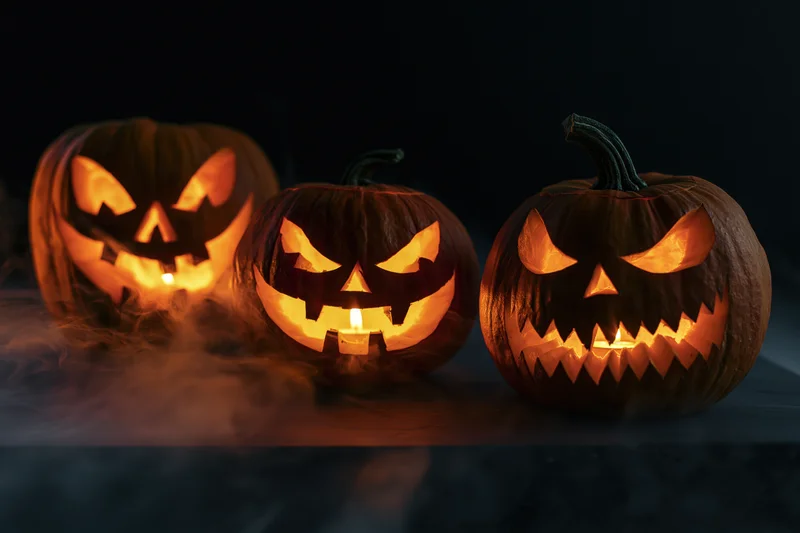 Three carved jack-o-lanterns glowing with candles inside on a dark surface with smoke swirling around them