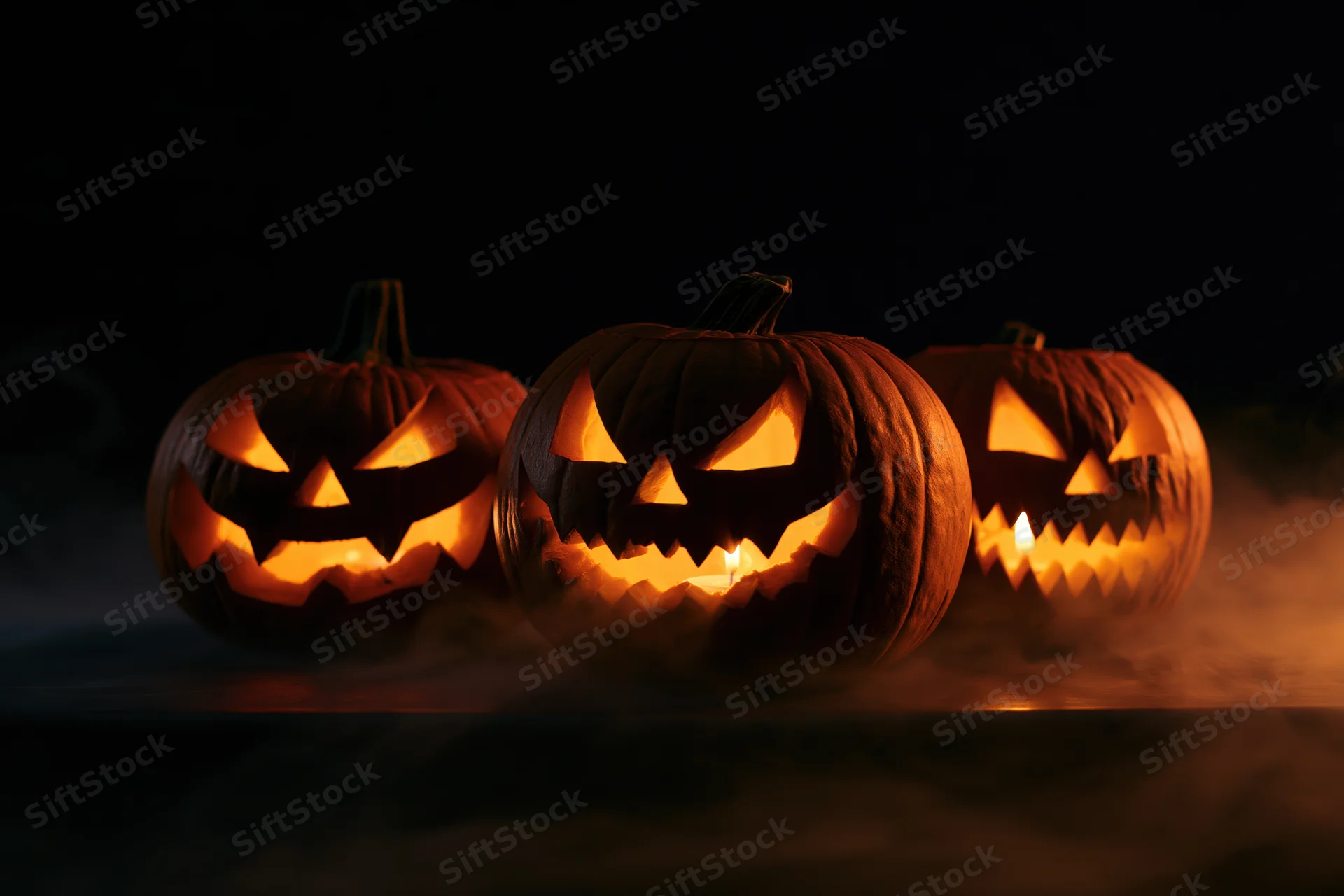 Three carved pumpkins glowing with candles inside on a dark smoky surface