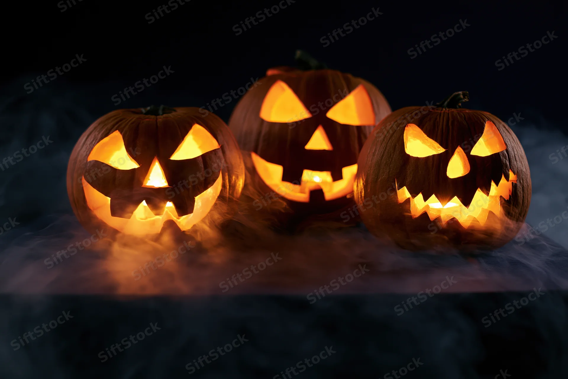 Three carved pumpkins glowing with light in a smoky dark setting
