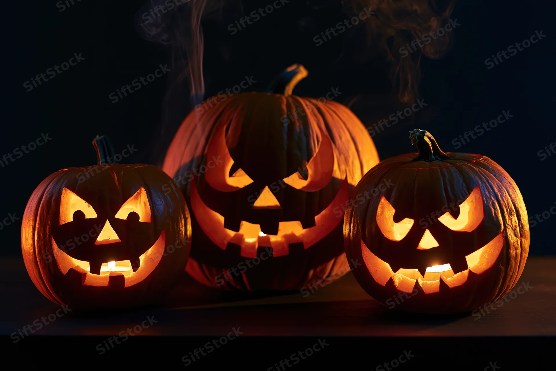 Three carved jack-o-lanterns glowing with candles and smoke on a dark surface