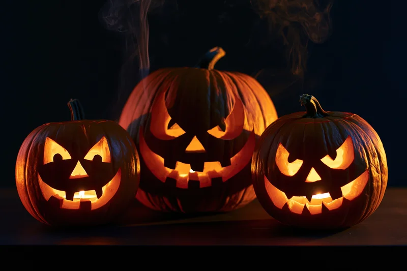 Three carved jack-o-lanterns glowing with candles and smoke on a dark surface
