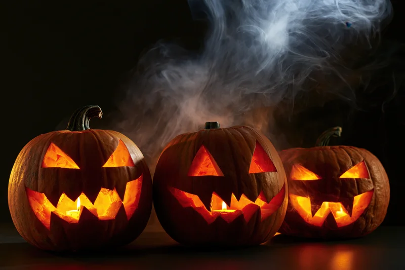 Three carved jack-o-lanterns glowing with candles and smoke on a dark surface