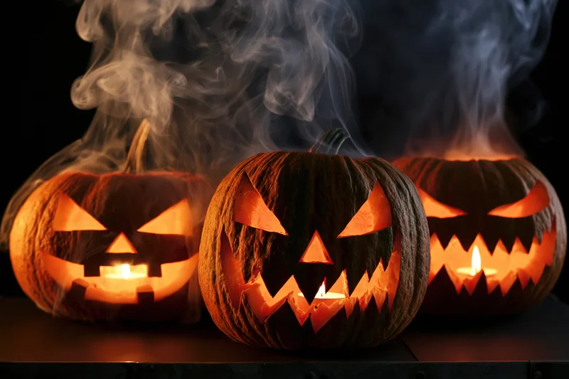 Three carved jack-o-lanterns glowing with candles and emitting smoke on a dark surface