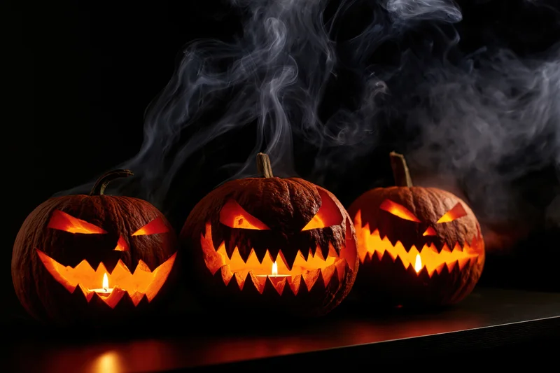 Three carved jack-o-lanterns glowing with candles and smoke on a dark surface