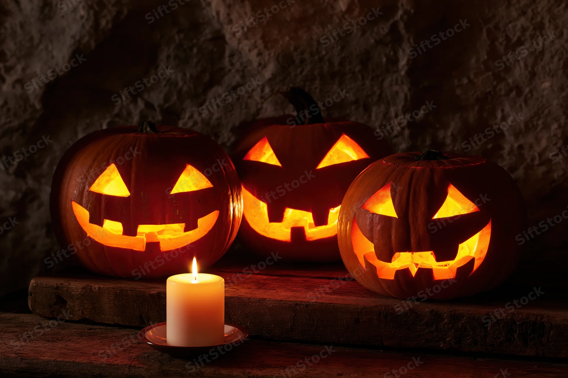 Three carved jack-o-lanterns glowing with candlelight on a wooden surface against a stone wall
