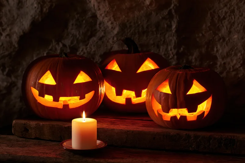 Three carved jack-o-lanterns glowing with candlelight on a wooden surface against a stone wall