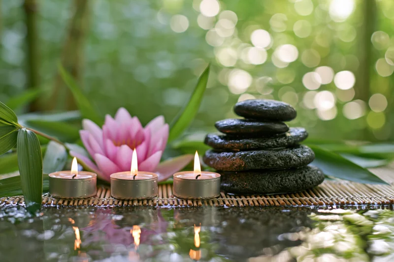 Three lit tea light candles beside a pink water lily and stacked stones on a bamboo mat over water
