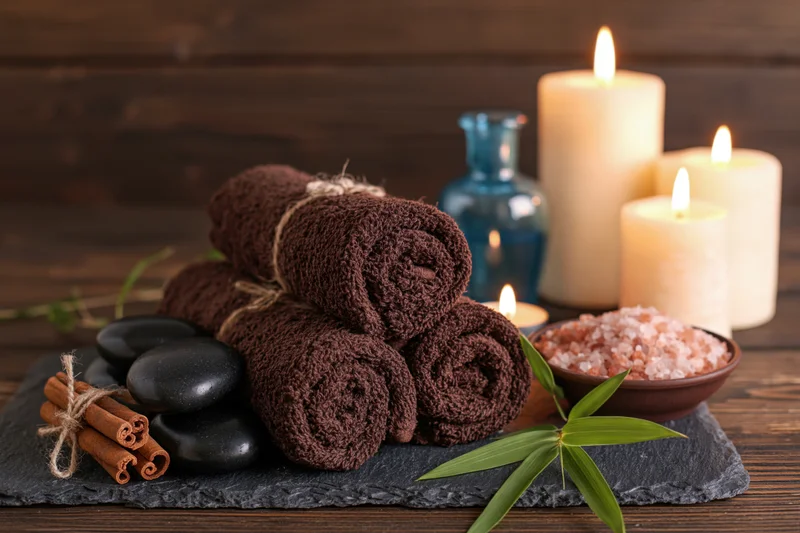 Rolled brown spa towels arranged on slate with candles, stones, salt, and cinnamon sticks