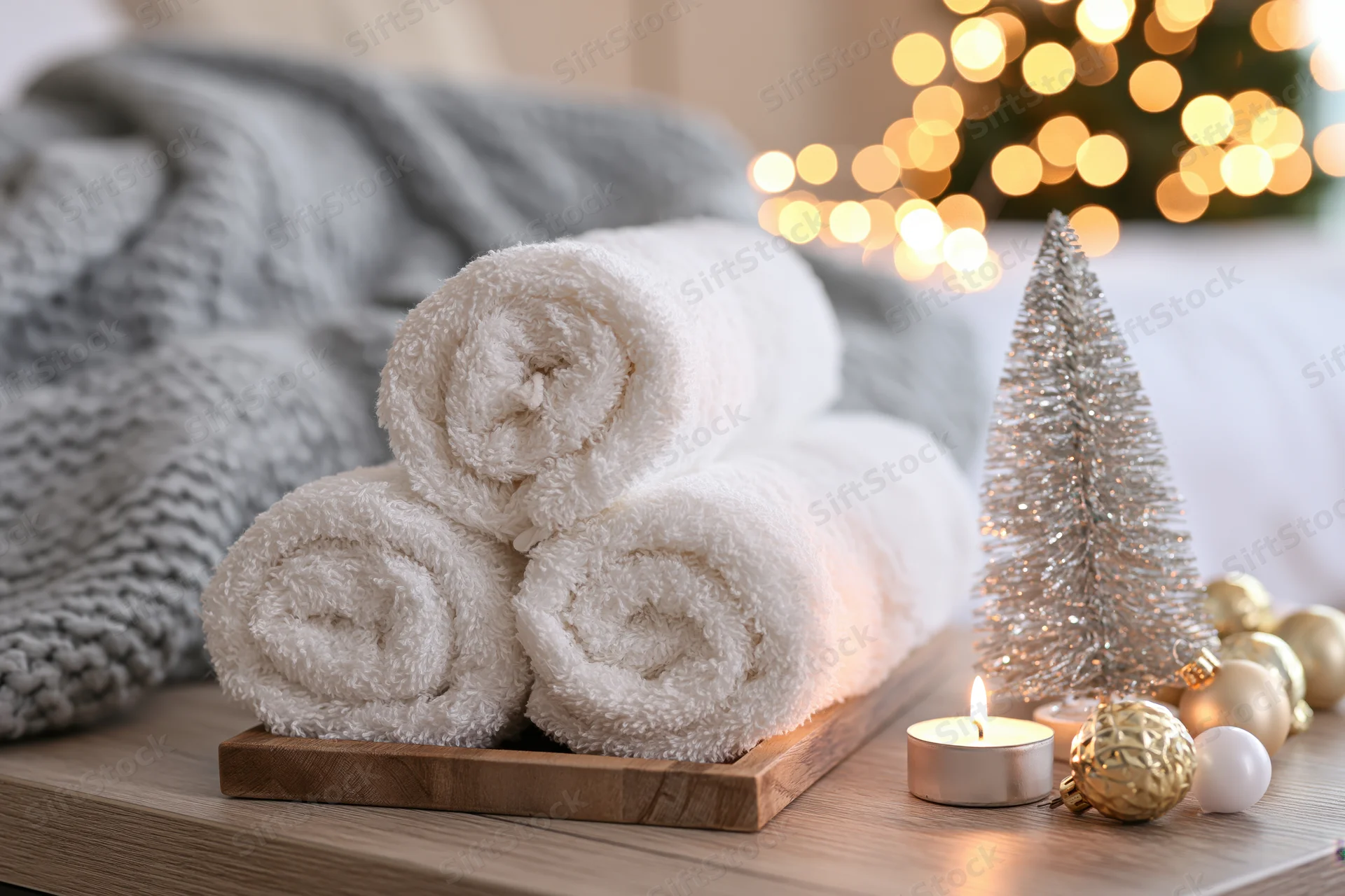 Three rolled white cotton towels stacked on a wooden tray beside a lit candle and christmas decorations on a table