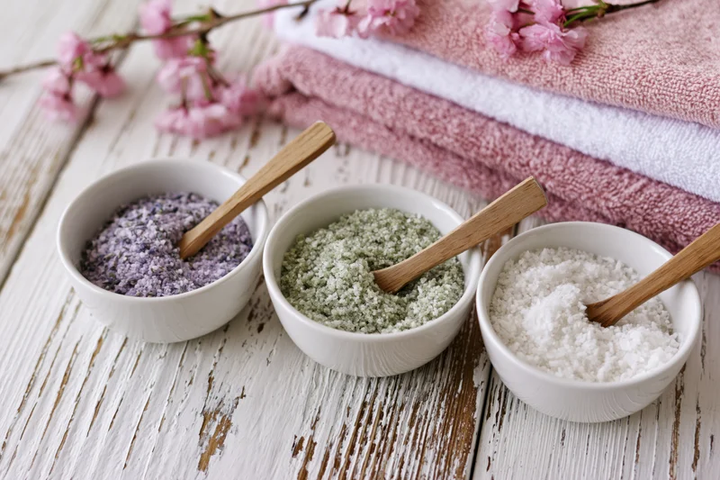 Three ceramic bowls with lavender, eucalyptus and coconut bath salts on a whitewashed wooden table beside pink towels and cherry blossoms