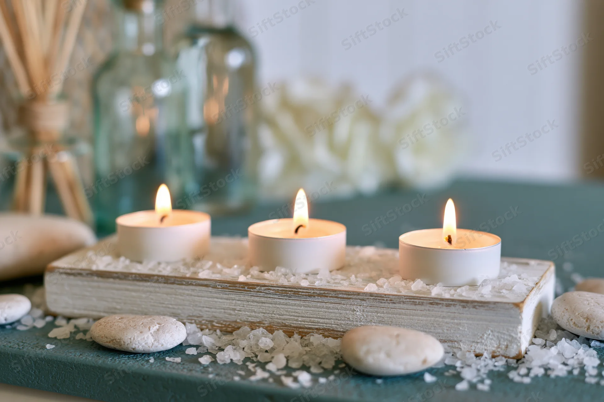 Three burning white tealight candles on a whitewashed wooden tray with salt and stones, surrounded by glass bottles and flowers on a teal surface