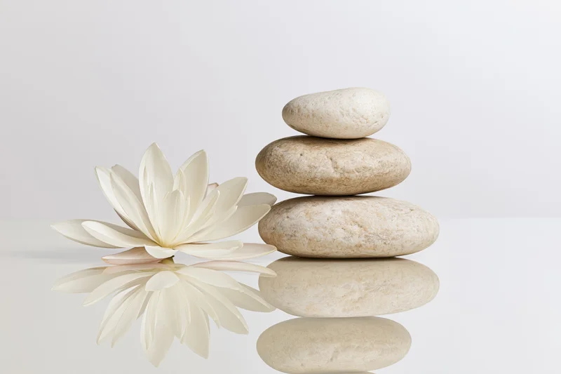 Three smooth river stones stacked in a balanced cairn beside a white lotus flower on a reflective white surface