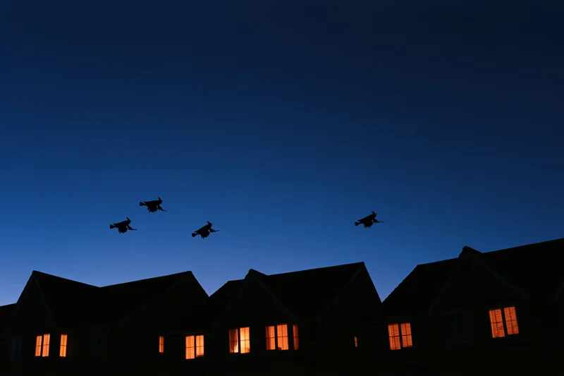 Witch silhouettes flying on broomsticks above suburban houses at twilight