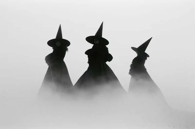 Three women in witch costumes standing in fog, silhouetted against misty background
