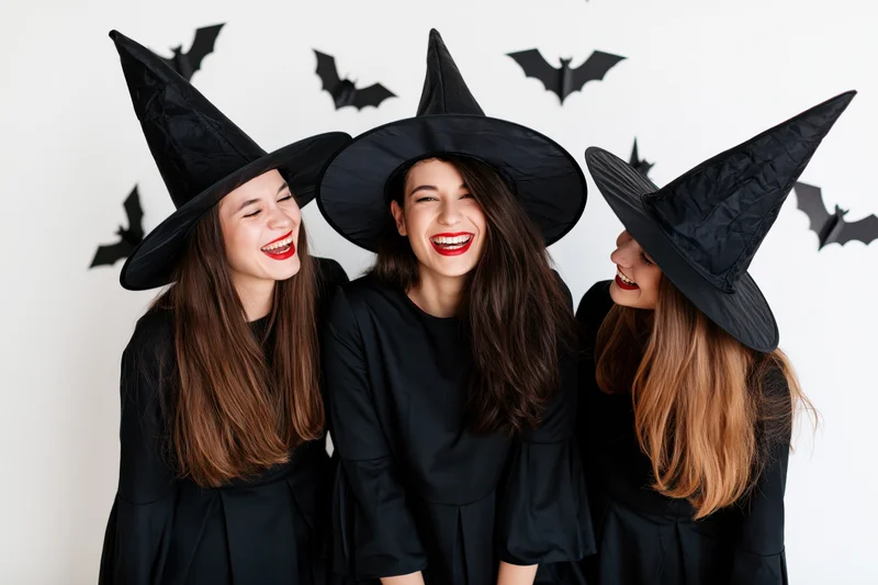 Three women in witch costumes laughing together indoors with bat decorations on the wall