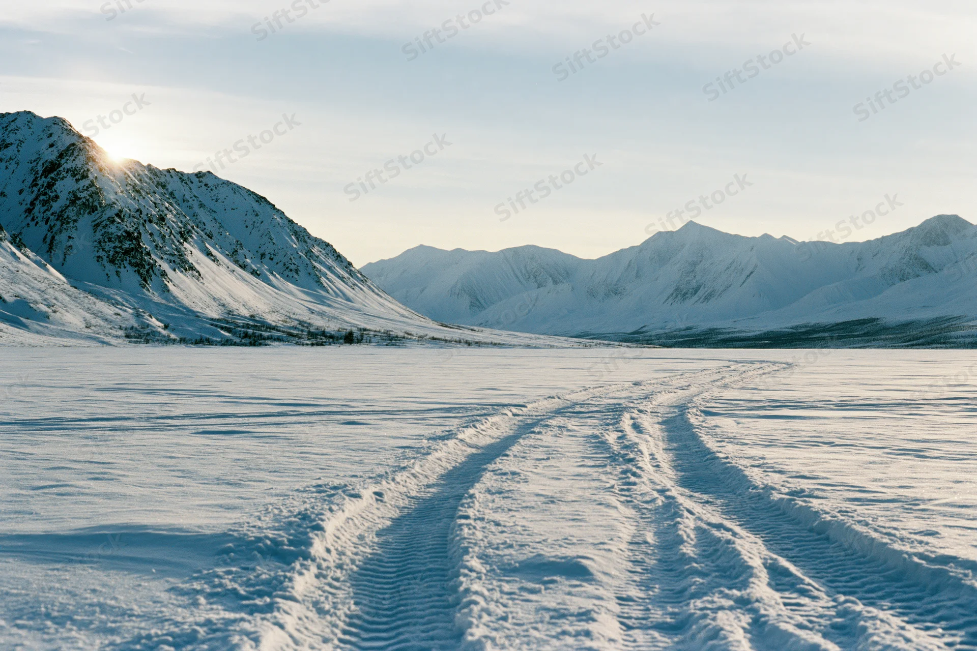 Tire tracks crossing a frozen snow plain leading toward snow-covered mountains under sunlight