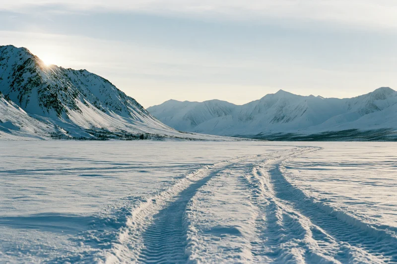 Tire tracks crossing a frozen snow plain leading toward snow-covered mountains under sunlight