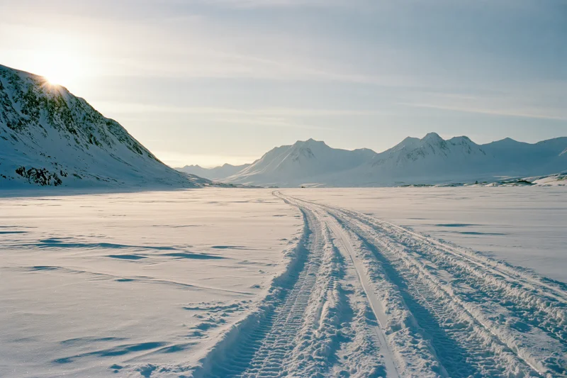 Tire tracks crossing a frozen snow plain toward distant snow-covered mountains under a pale sky at sunrise