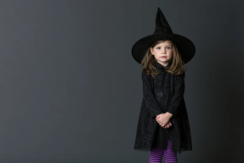 Toddler girl in witch costume standing and looking directly at camera in studio setting