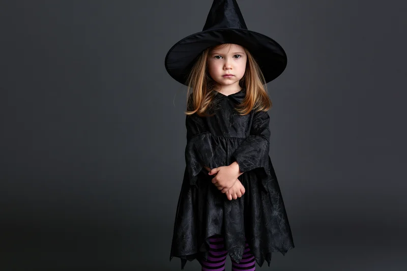 Toddler girl in witch costume standing and looking directly at camera in studio setting