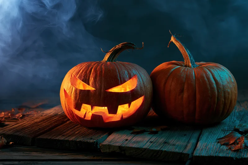 Carved pumpkin glowing with orange light beside a whole pumpkin on weathered wooden planks with smoke in dark background