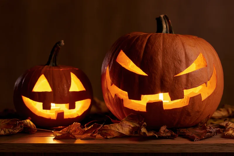 Two carved pumpkins glowing with candles on a wooden table surrounded by dry leaves