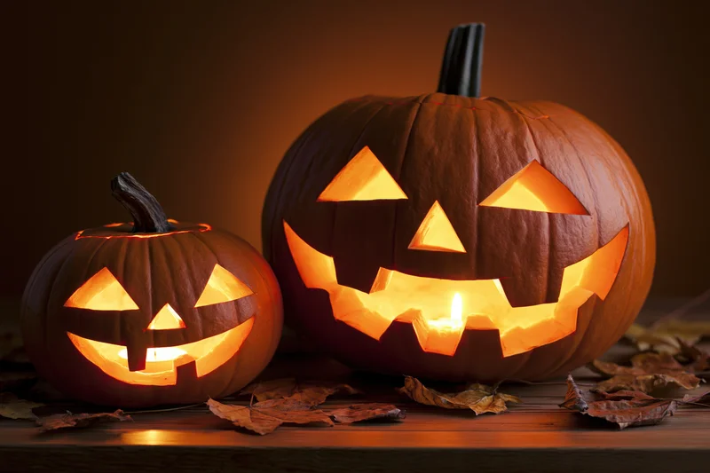 Two carved pumpkins glowing with candles on a wooden table surrounded by dry leaves