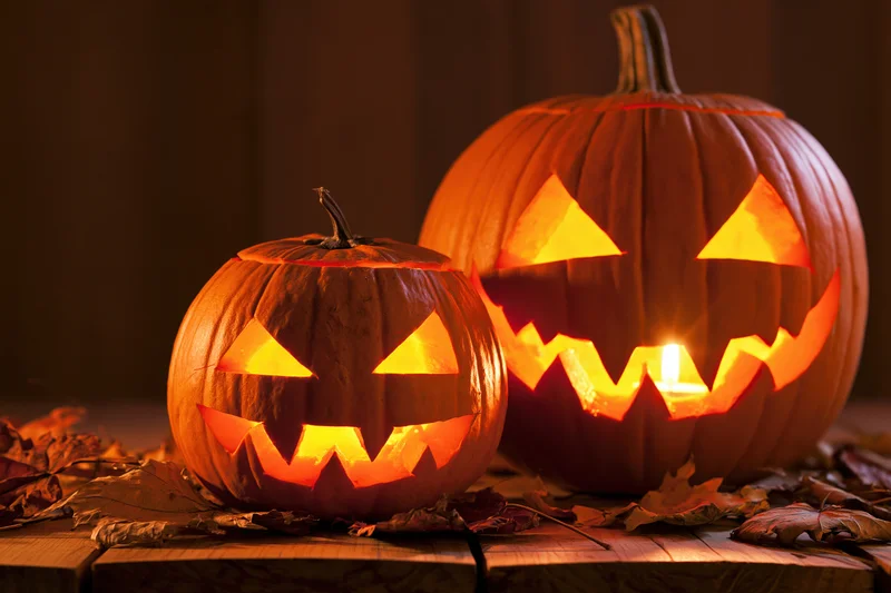 Two carved pumpkins glowing with candles on a wooden table surrounded by dry leaves