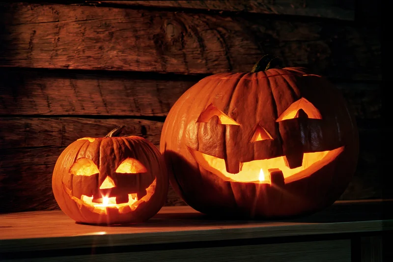 Two carved pumpkins glowing with candles on a wooden table against a rustic wall