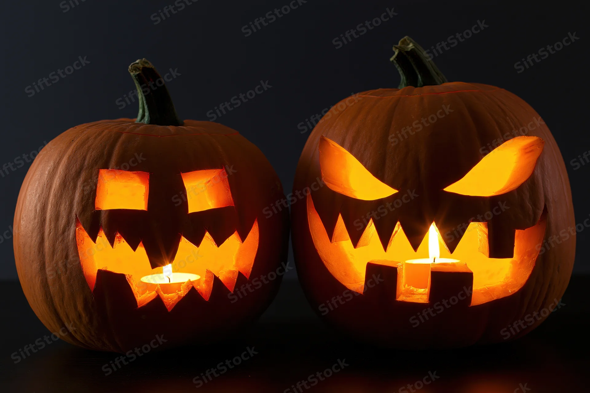 Two carved pumpkins with lit candles inside, side by side on a dark surface