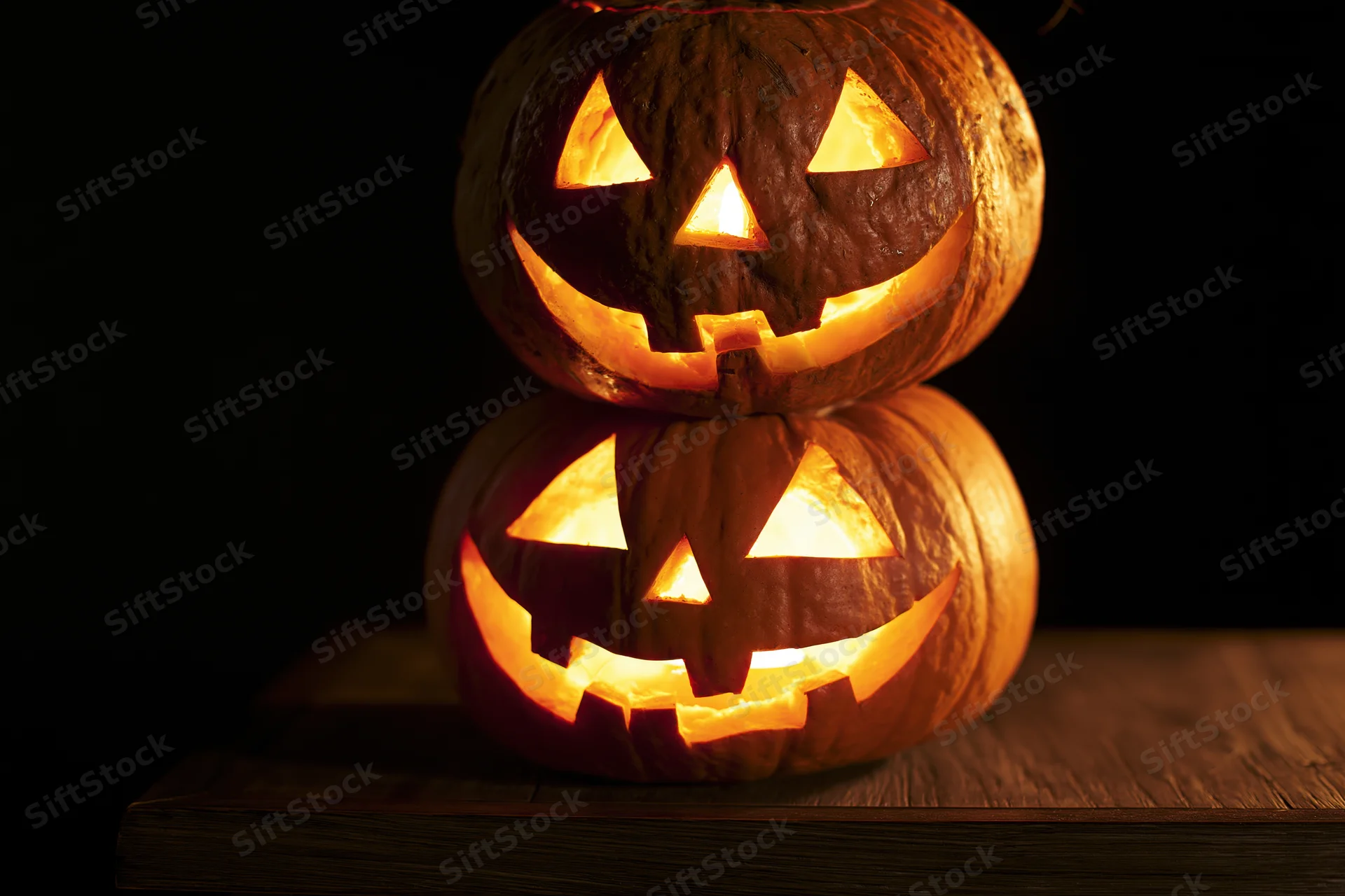 Two carved jack-o-lanterns stacked on a wooden table glowing in the dark