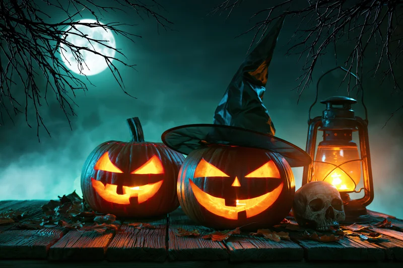 Two carved jack-o-lanterns with glowing orange faces on a wooden table under a full moon at night, surrounded by a witch hat, lantern, skull, and bare branches
