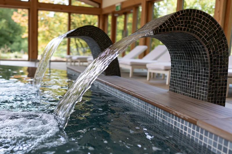 Curved mosaic water jets cascading into indoor pool with lounge chairs and wooden structure
