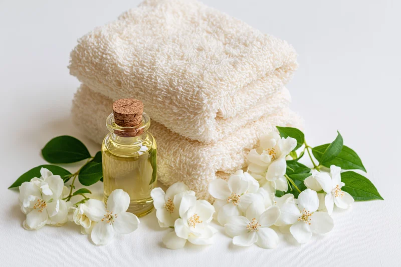 Folded cream towels with jasmine flowers and essential oil bottle on white surface
