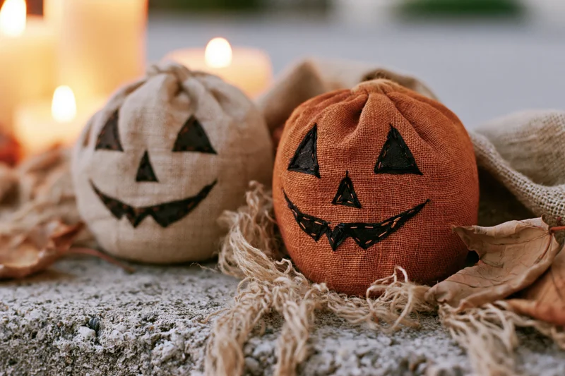 Two handmade fabric jack-o-lanterns with stitched faces resting on a rough concrete surface surrounded by dried leaves and glowing candles