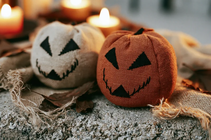 Two handmade fabric jack-o-lanterns with stitched faces resting on rough concrete beside lit candles and dried leaves