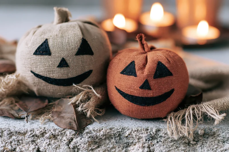 Two handmade fabric jack-o-lanterns with stitched faces resting on a rough concrete surface surrounded by lit candles and dried leaves