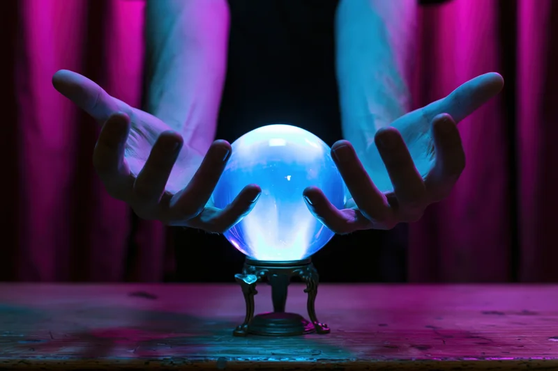 Hands hovering over a glowing blue crystal ball on a wooden table with purple curtains in the background