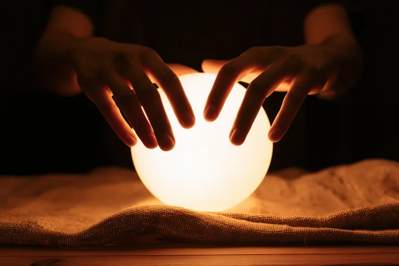 Hands hovering over a glowing crystal ball on a burlap cloth indoors