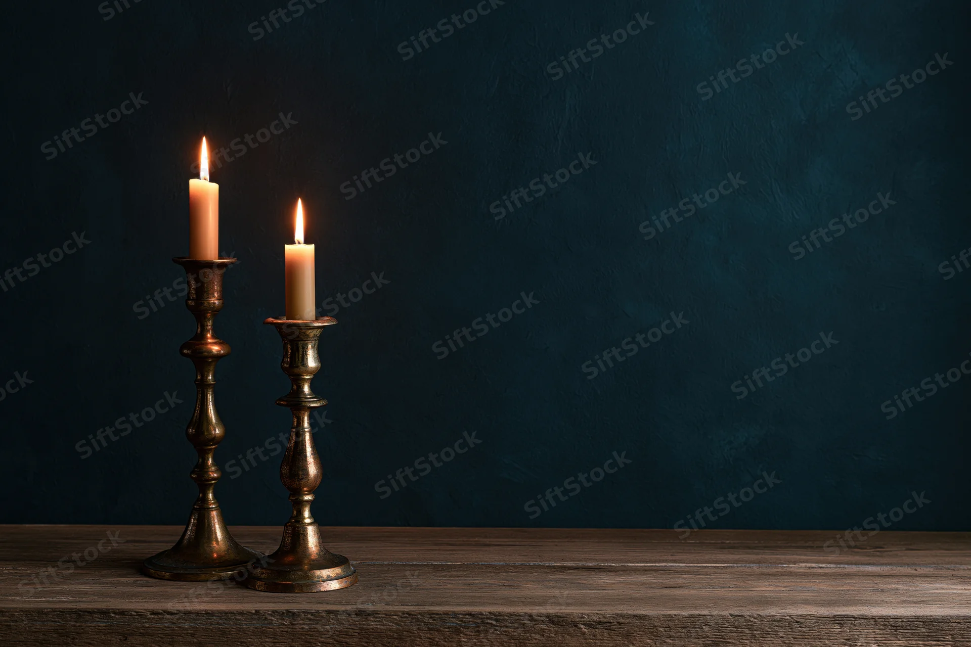 Two lit candles in antique brass candlesticks on a wooden table against a dark blue textured wall