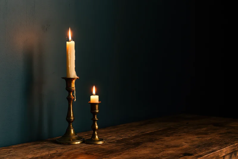 Two lit candles in brass candlesticks on a wooden table against a dark wall