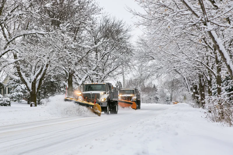 Two municipal snowplow trucks clearing snow from a residential street lined with snow-covered trees