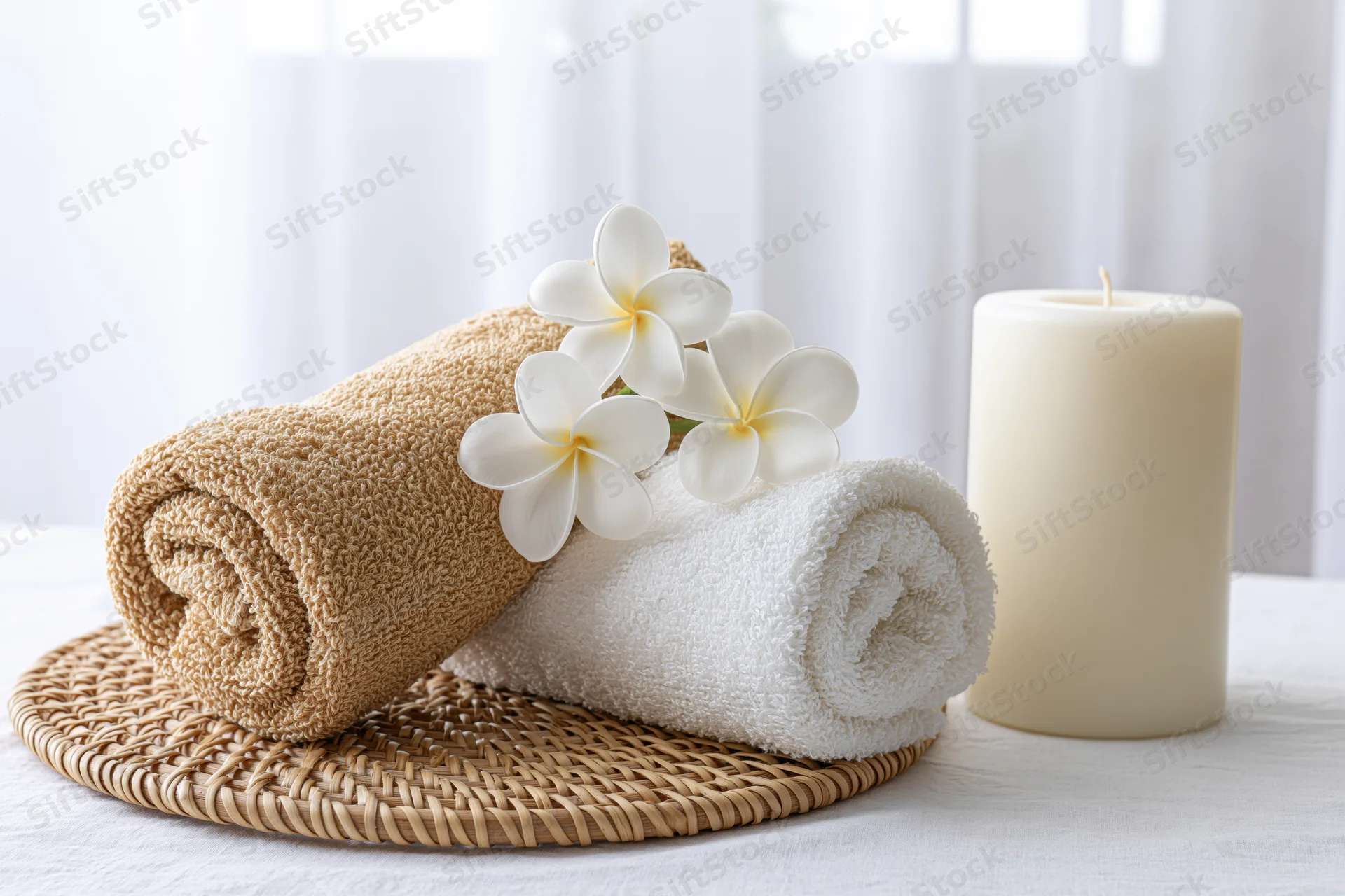 Two rolled spa towels with frangipani flowers and a candle on a woven trivet on a white table